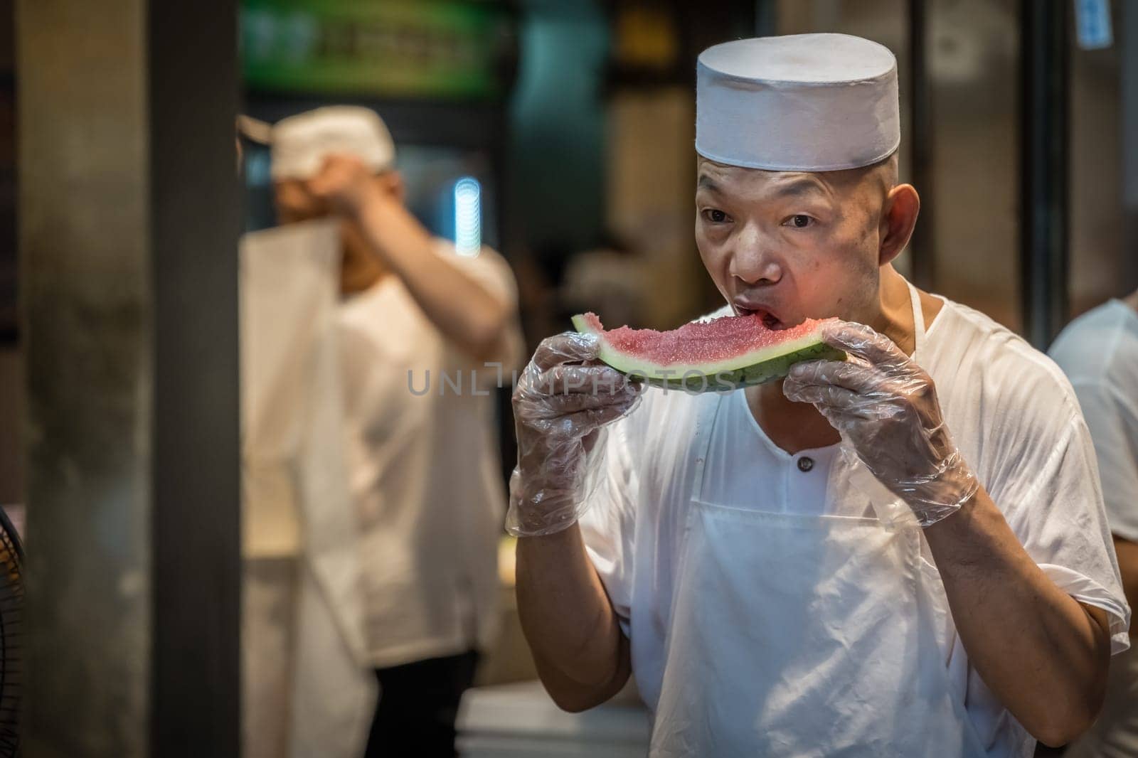 Xian, China -  July 2019 : Chinese chef taking his worki break and eating delicious and ripe slice of watermelon on the street in the Muslim Quarter