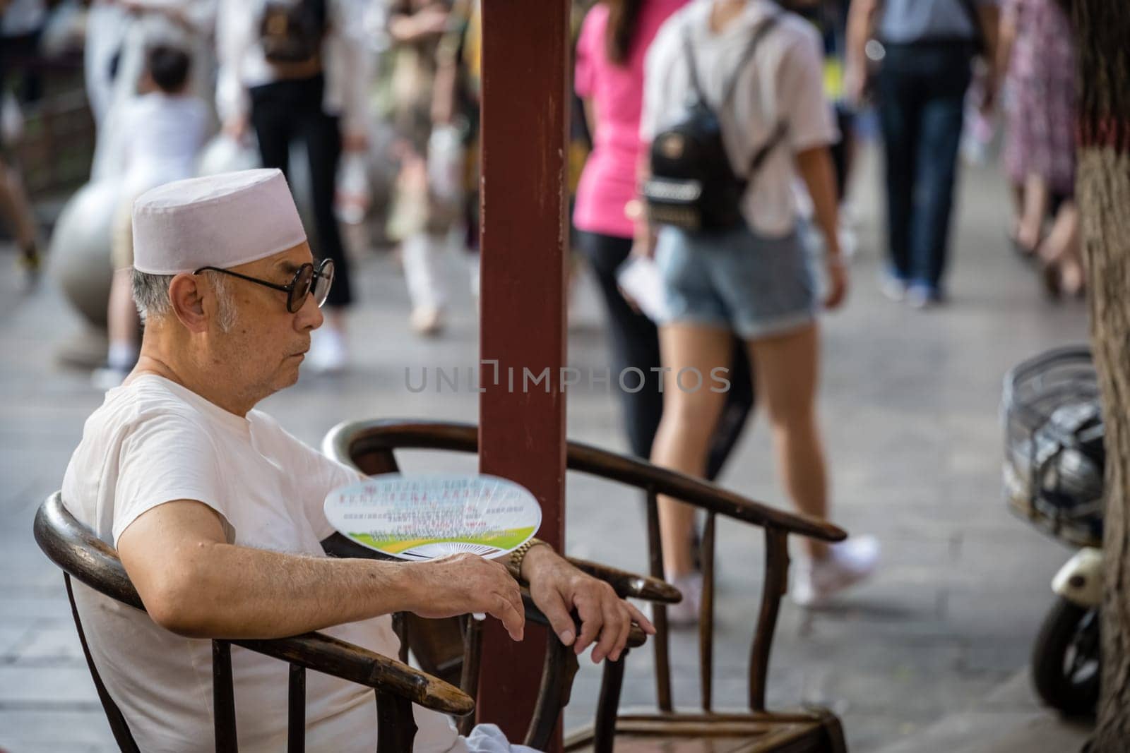 Xian, China -  July 2019 : Tired Chinese man resting on a chair and cooling himself by waving with a small fan on a hot day on the street in the Muslim Quarter