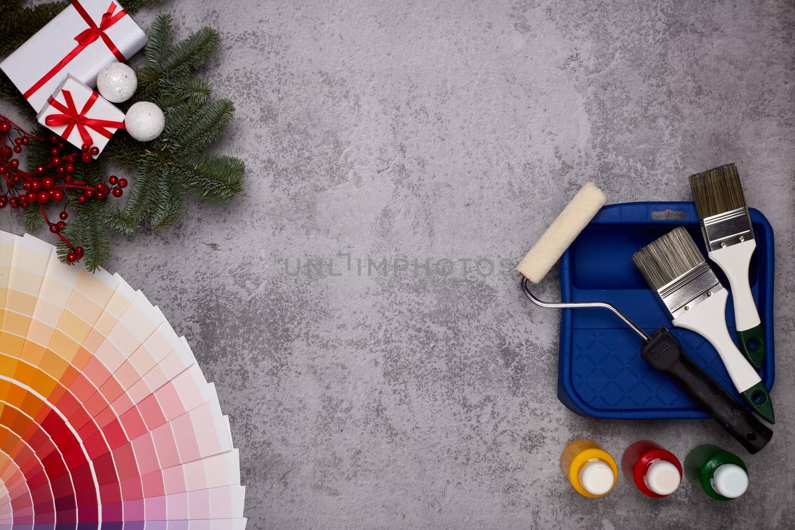 Color palette guide, paint brushes, roller, cans of paint, fir tree branch and gift boxes on grey background. Top view, copy space, New Year and Christmas. Concept of construction or design office