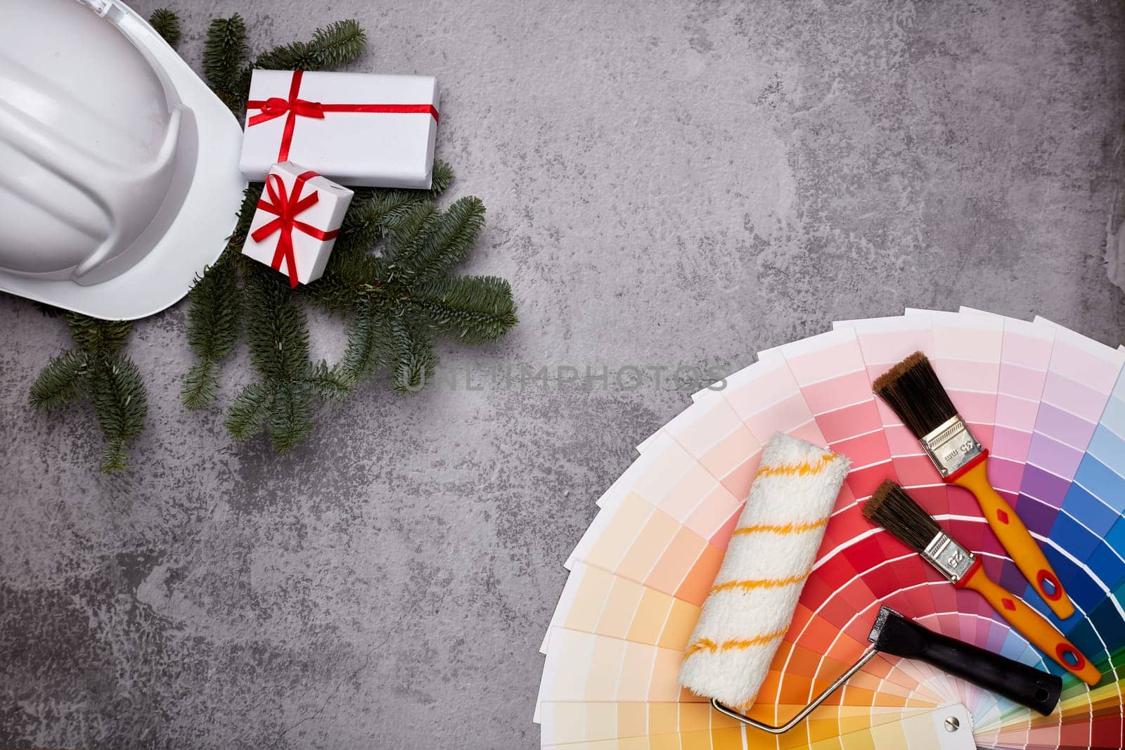 Construction hard hat, color palette guide, paint brushes and roller, fir tree branches and gift boxes on grey background. Top view with copy space, New Year and Christmas. Concept of construction or design office