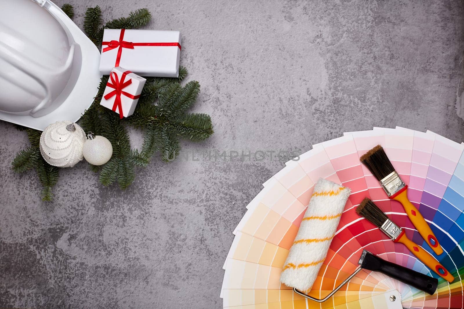 Construction hard hat, color palette guide, paint brushes and roller, fir tree branches and gift boxes on grey background. Top view with copy space, New Year and Christmas. Concept of construction or design office