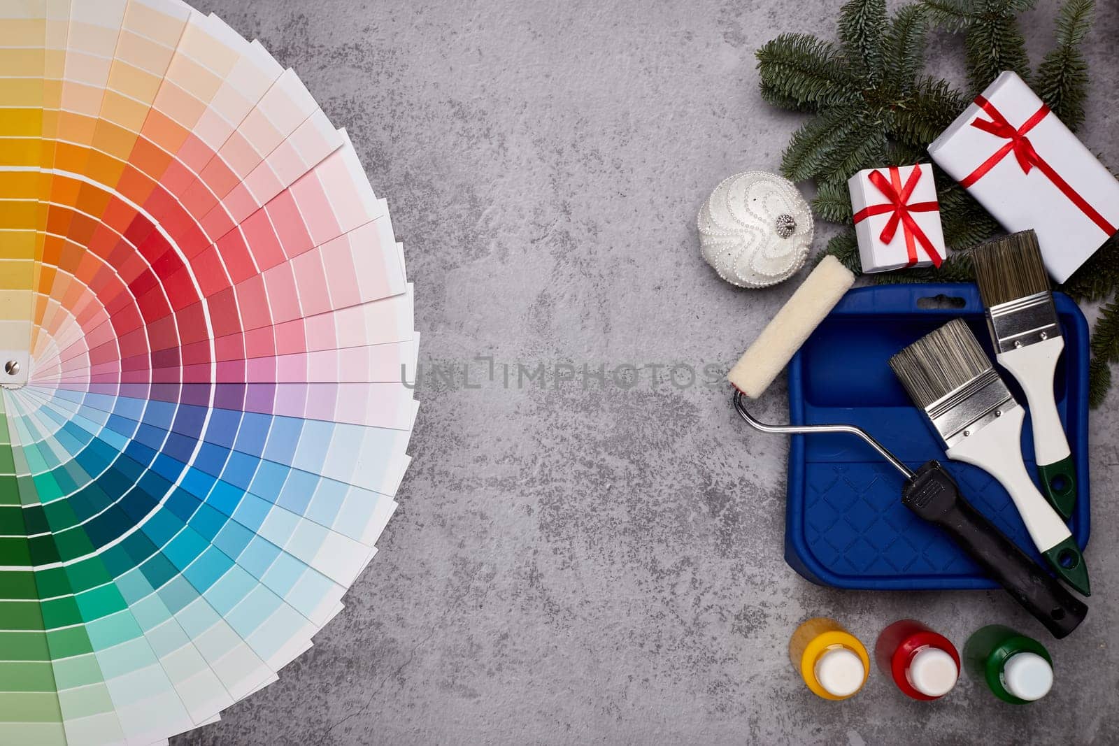 Color palette guide, paint brushes, roller, cans of paint, fir tree branch and gift boxes on grey background. Top view, copy space, New Year and Christmas. Concept of construction or design office