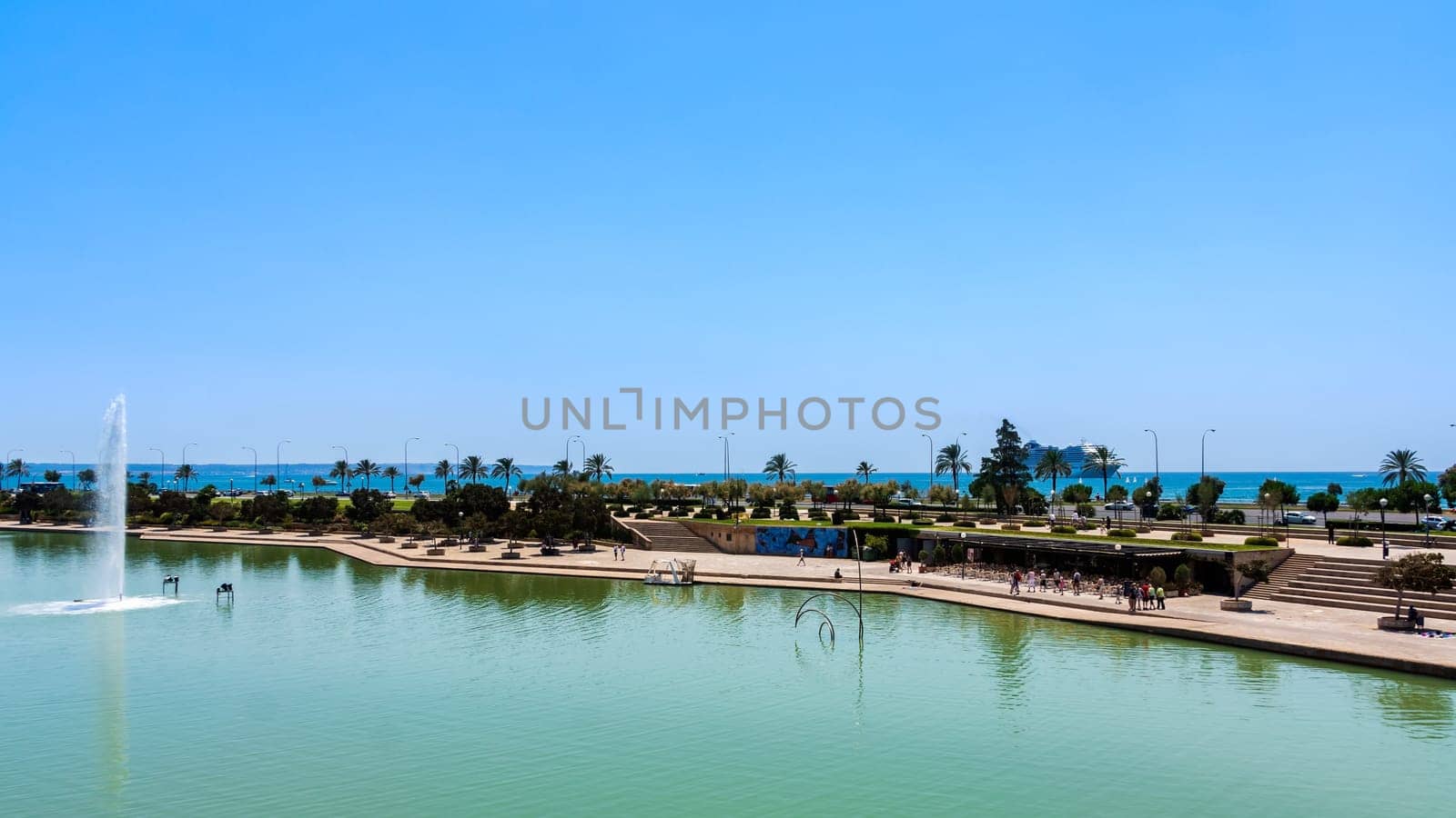Tourists walking on promenade between The Cathedral of Santa Maria of Palma in Mallorca with fountain and pond in front and Balearic Sea in background