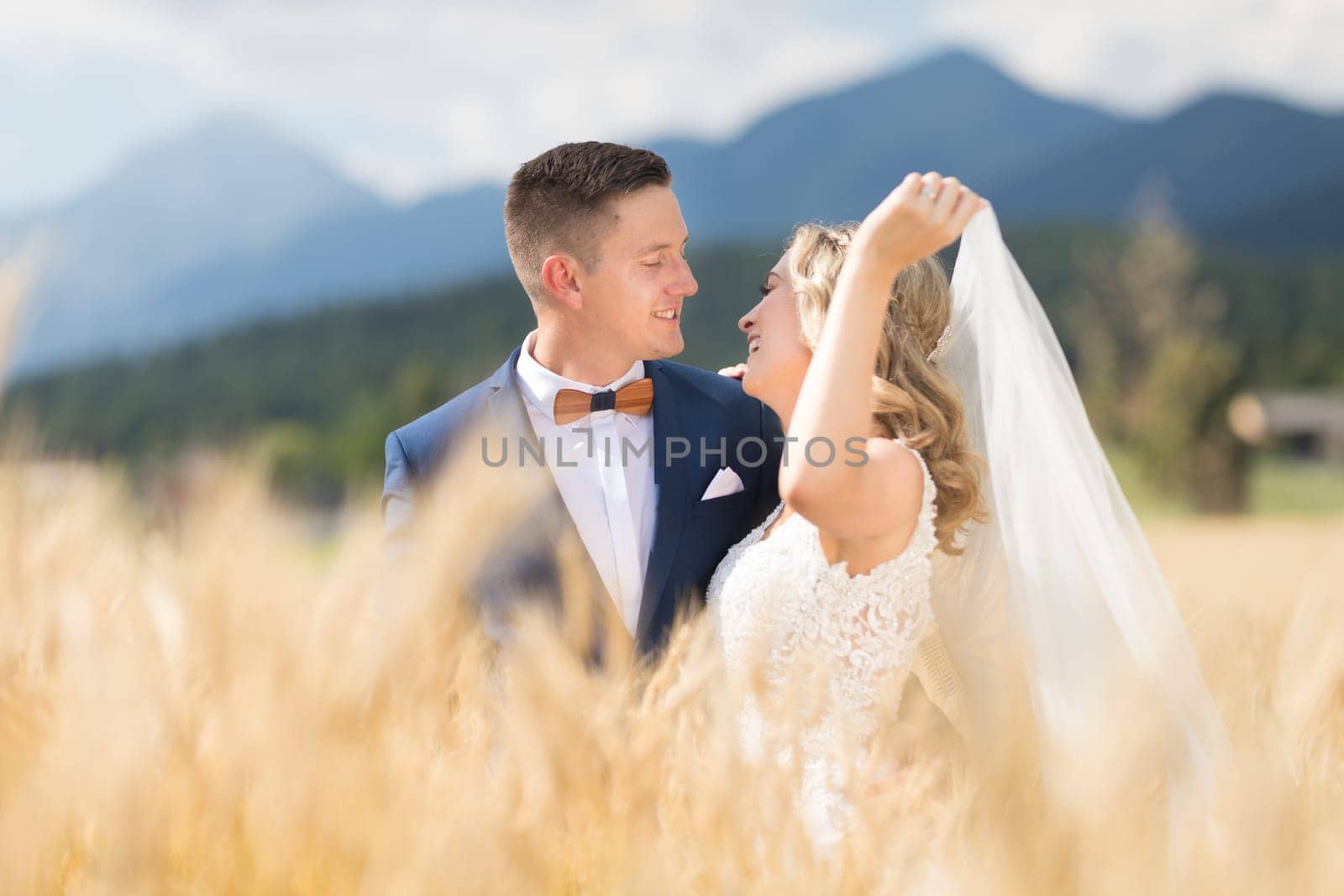 Groom hugs bride tenderly while wind blows her veil in wheat field somewhere in Slovenian countryside. Caucasian happy romantic young couple celebrating their marriage.