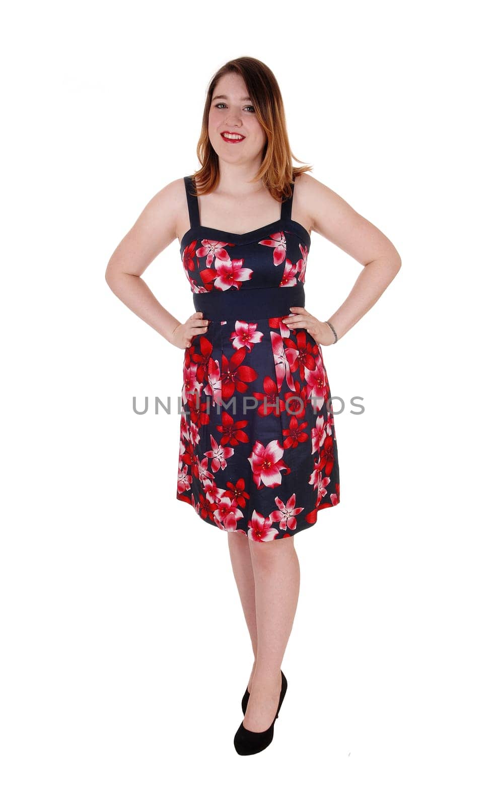 A beautiful young woman standing in black heels and a colorful.printed dress with her hands on her hips, isolated for white.background.