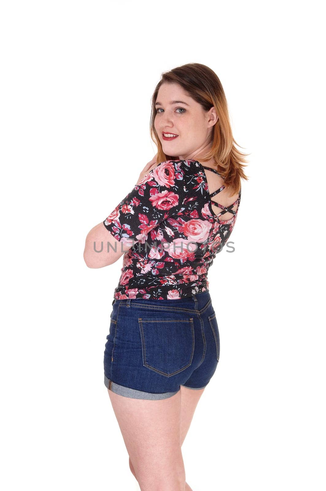 A beautiful young woman standing from the back in jeans shorts and.a colorful blouse with her arms crossed, isolated for white background.