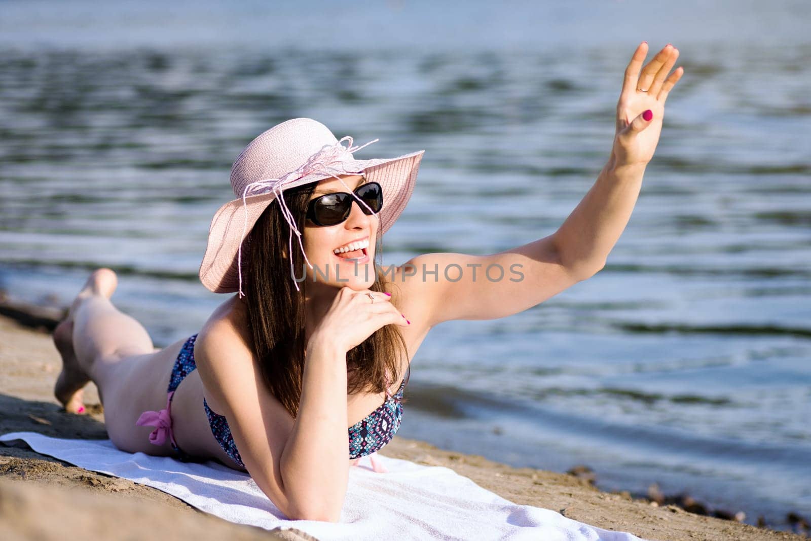 The smiling cheerful woman is greeting, saying hello and waving hand to someone on the beach at summer.