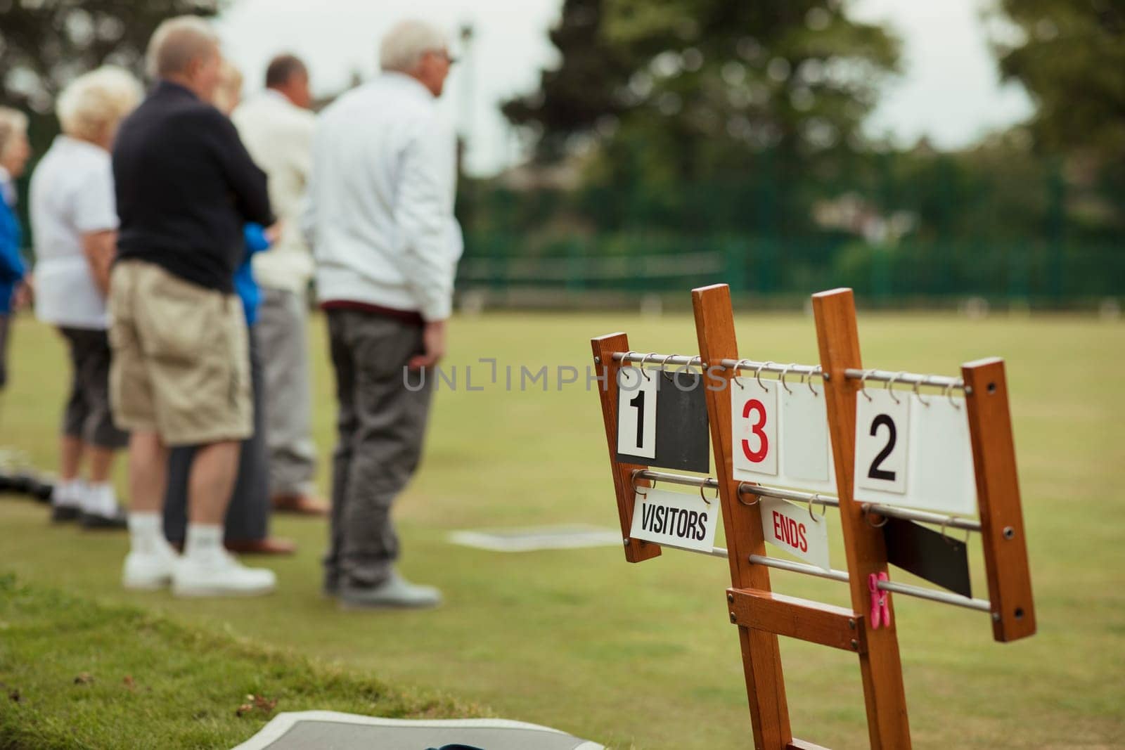 A close up shot of a lawn bowling scoreboard on grass, with senior adults spectating the lawn bowling game.