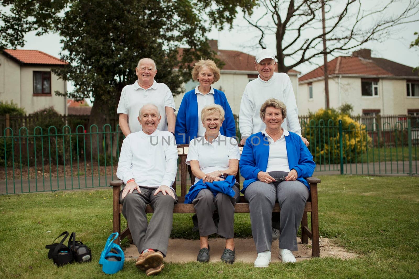A group of senior adults sitting on a bench at a bowling green, looking at the camera.