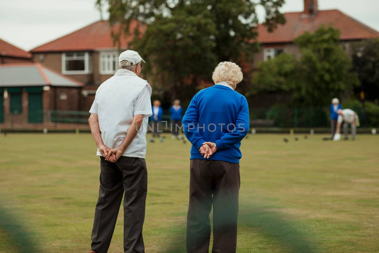 A rear view shot of a senior couple spectating a lawn bowling game, standing with their hands behind their back.