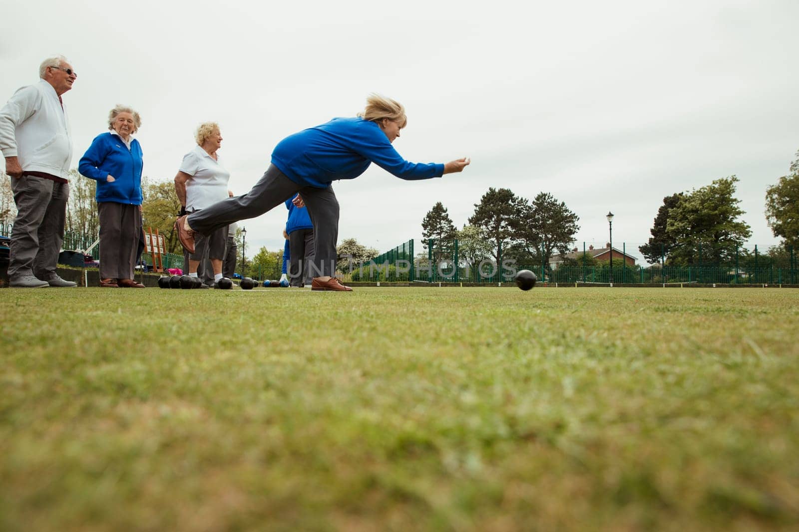 A side view shot of a group of senior friends lawn bowling on grass, with one woman who has taken her shot.