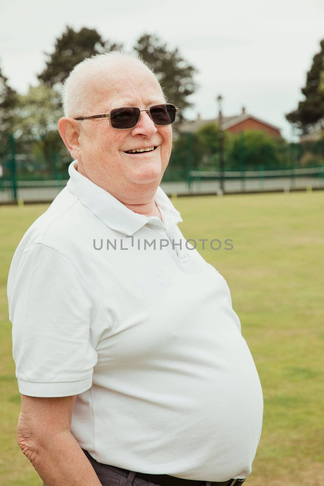 A portrait of a senior man smiling at a bowling green. He is wearing sunglasses and looks confident.