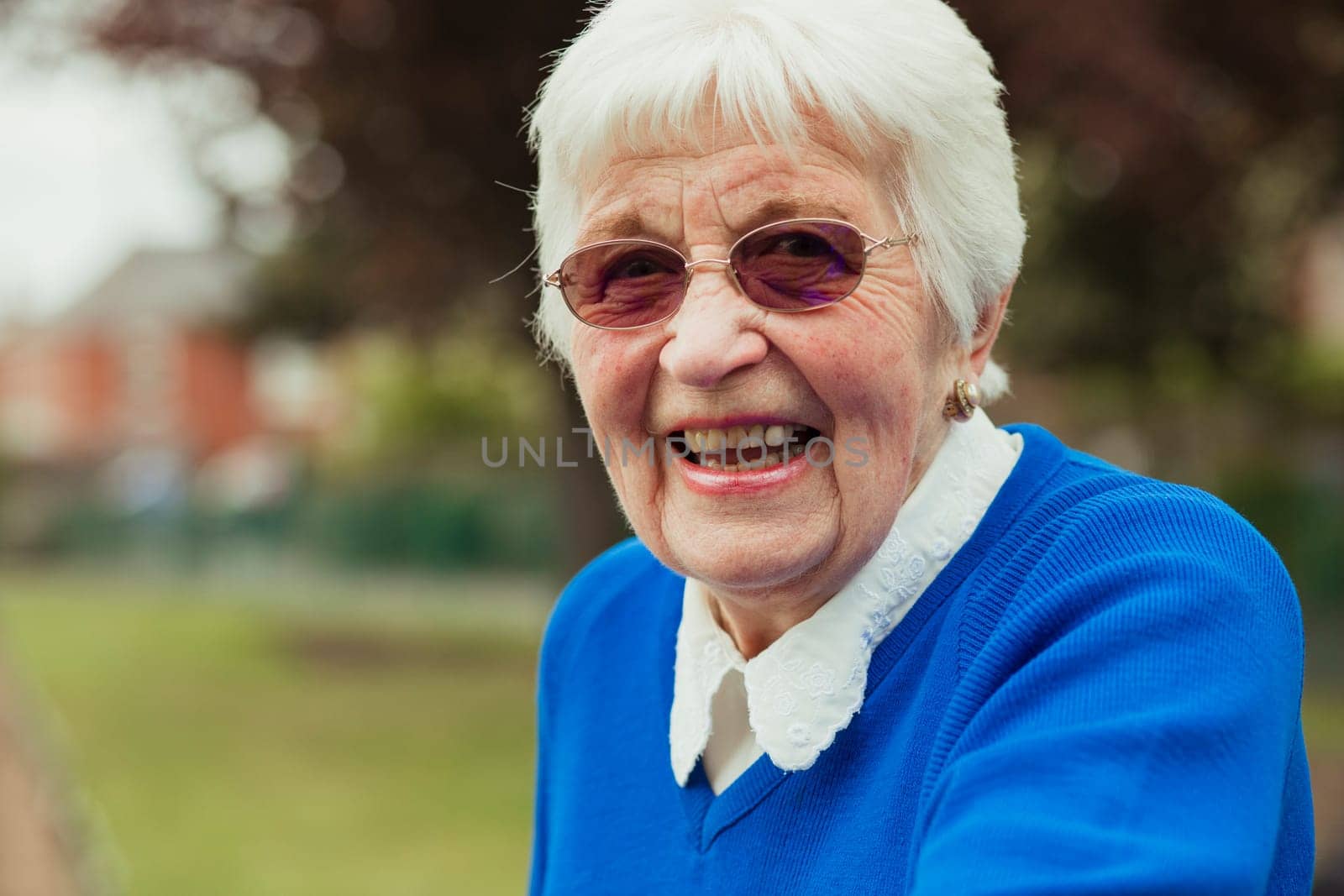 A close up shot of a senior woman smiling, looking at the camera. She is wearing rose coloured sunglasses.