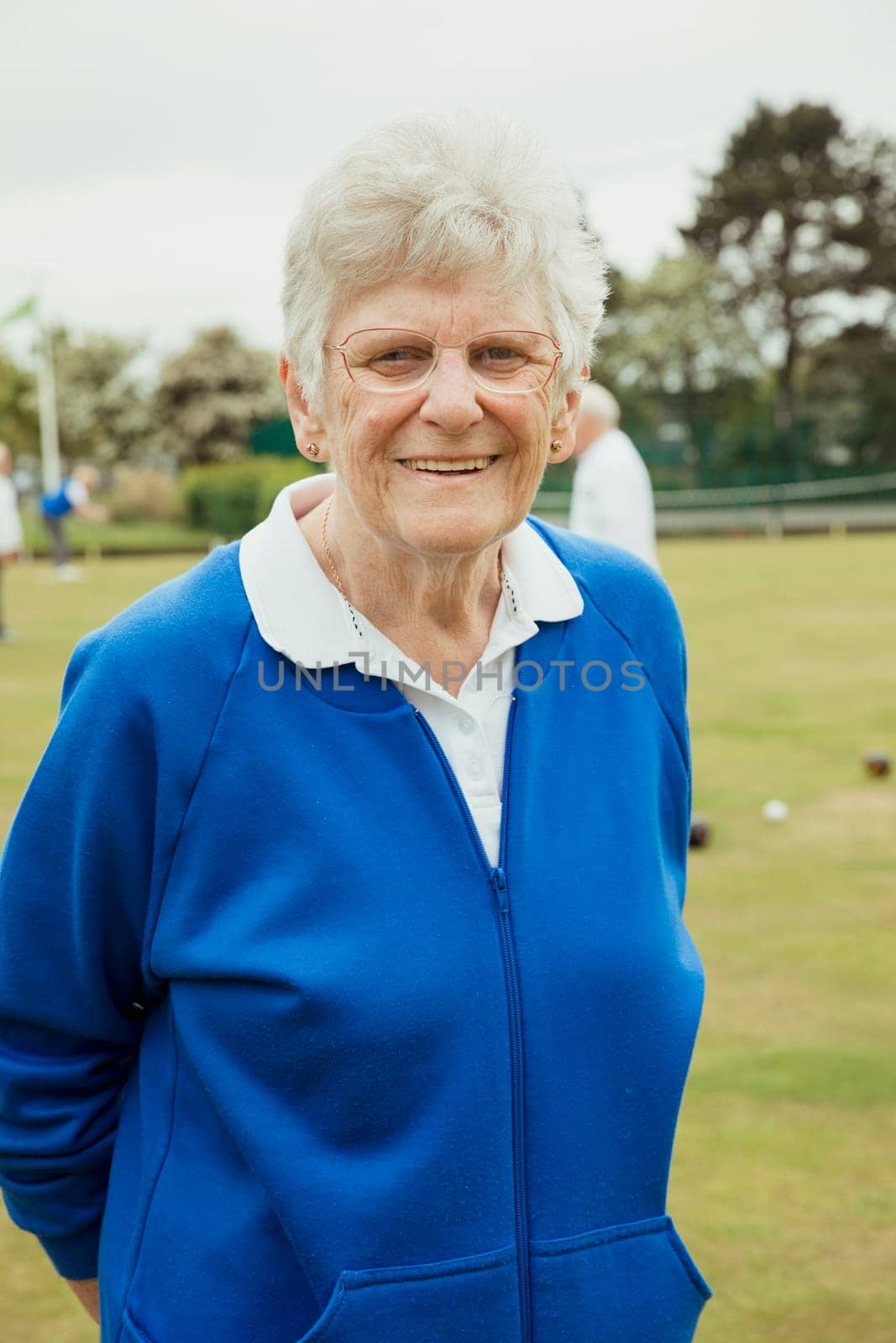 A portrait of a senior woman smiling at a bowling green. She is wearing glasses.