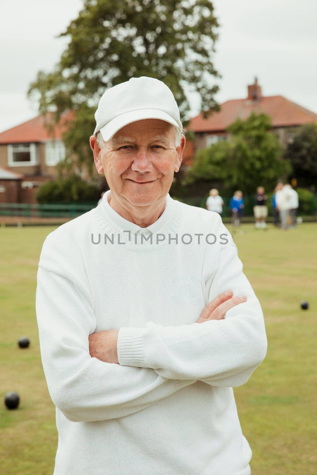 A portrait of a senior man smiling at a bowling green. He is wearing a white cap and is crossing his arms.