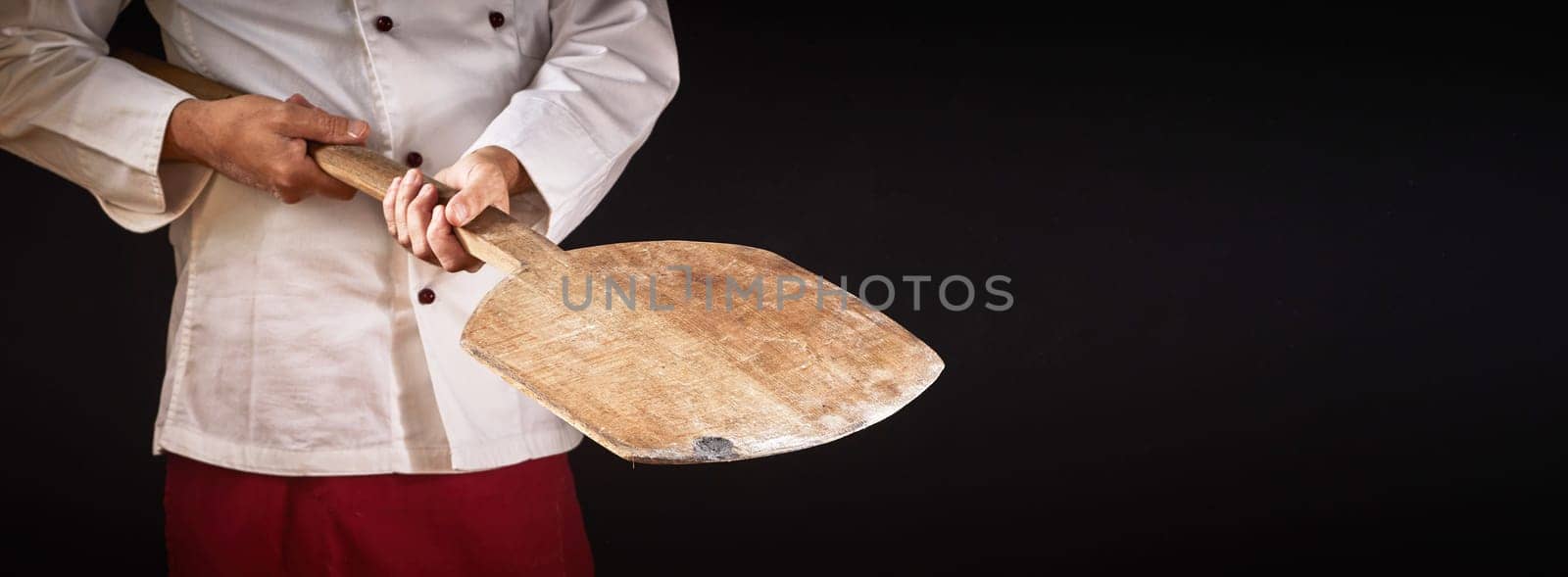 Chef holding an empty old wooden pizza paddle suitable for food placement over a black background with copy space in a panorama banner