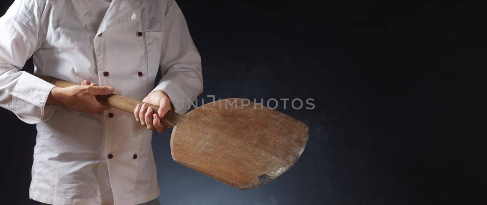 Chef in white uniform holding an empty wooden pizza paddle with placement copy space for food over a dark background