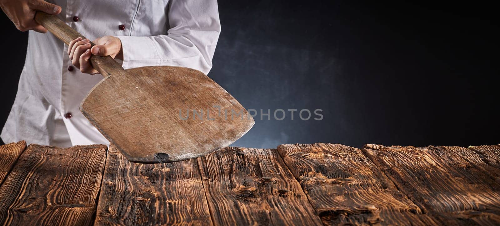 Chef holding an empty wooden pizza paddle over an old rustic kitchen table in a panorama banner with copy space for product or food placement