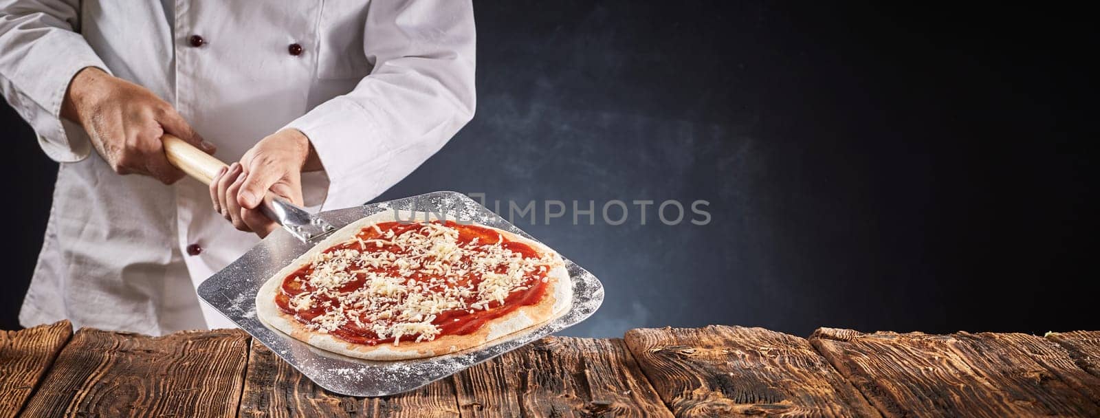 Chef making a traditional Italian pizza holding the crust with tomato paste and grated cheese on a paddle over a rustic table in a panorama banner with copy space
