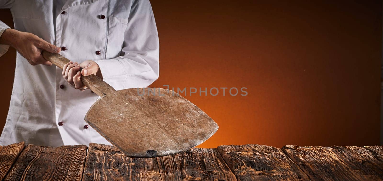 Restaurant chef in a pizzeria holding a wooden paddle over a rustic table against a brown background with copy space in a wide angle panorama with glow of the oven fire