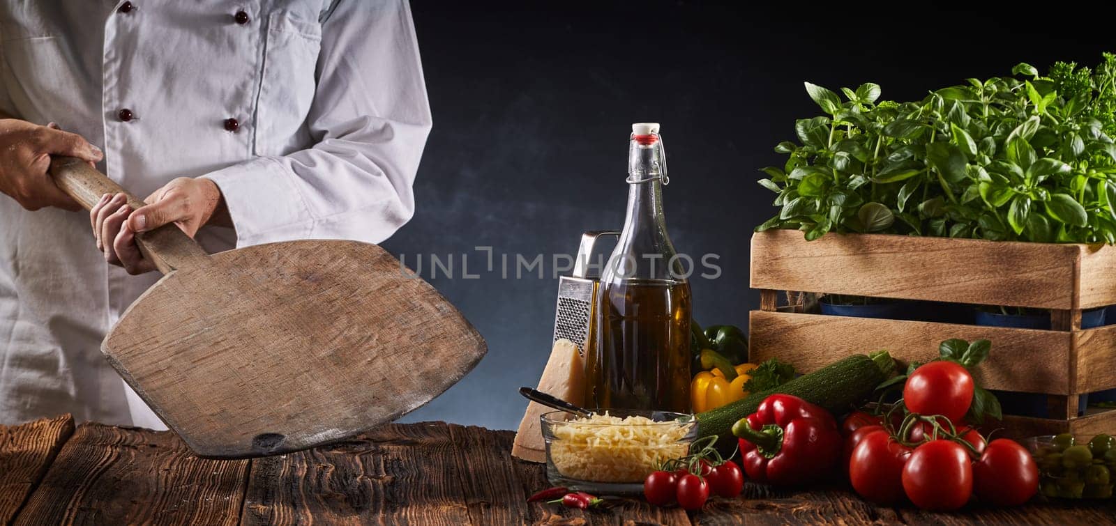 Chef holding a wooden paddle in a pizzeria above a rustic table with assorted fresh ingredients for Italian cuisine in a panorama banner