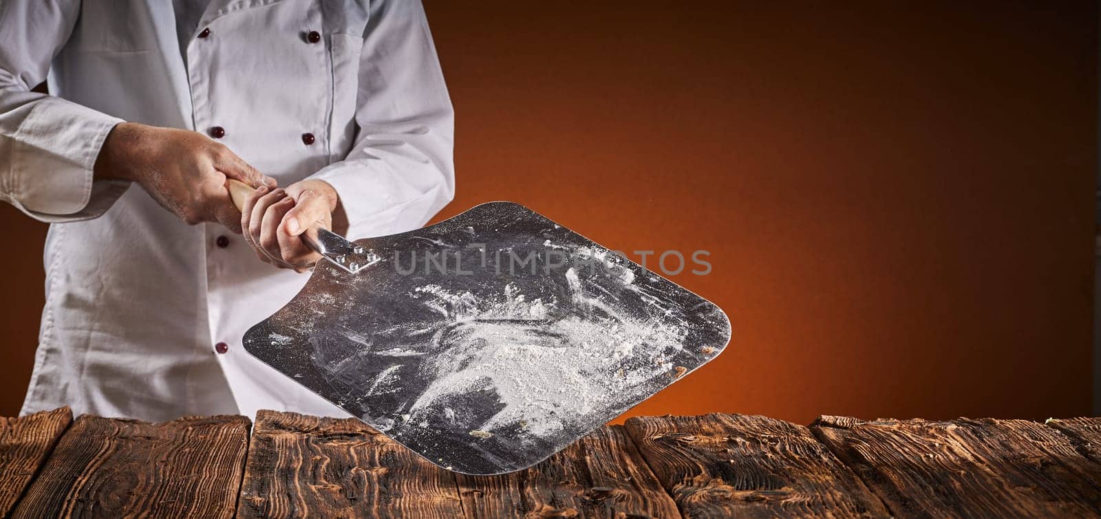 Pizza chef holding a floured metal paddle above a rustic wooden table in the kitchen over a brown background with copy space in a wide angle panorama with glow of the oven fire for food placement