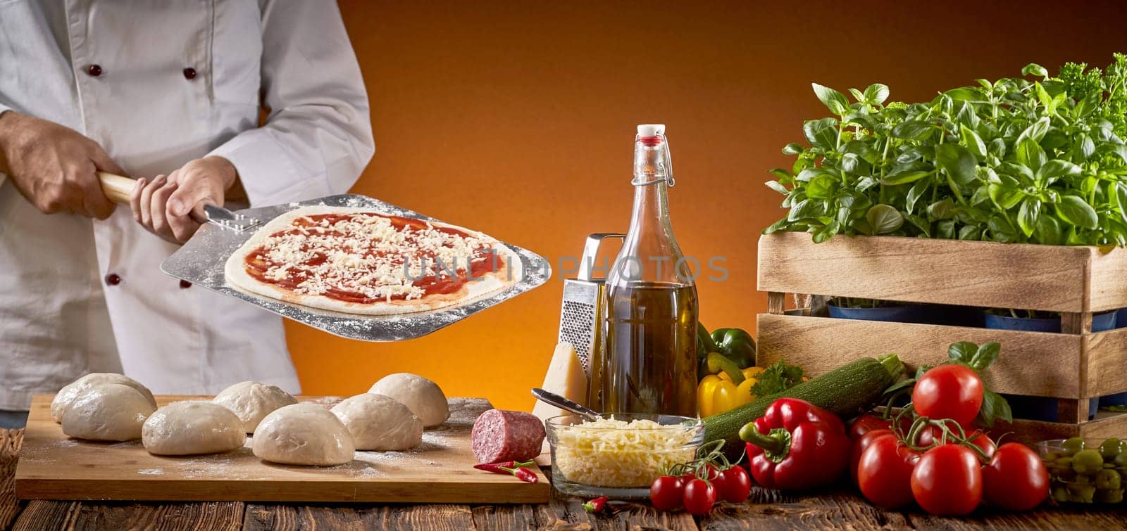 Cook preparing an Italian pizza in a restaurant holding a paddle with pastry base behind a rustic table with assorted fresh ingredients and portions of dough in a panorama banner