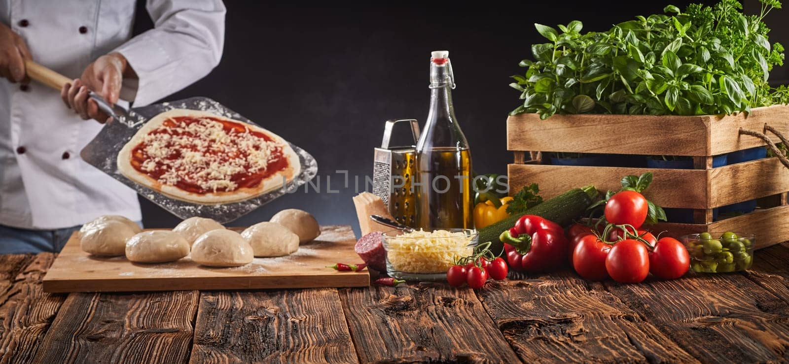 Chef in a pizzeria preparing an Italian pizza holding a paddle with a pastry base, tomato and cheese behind a rustic table with fresh ingredients and raw dough in a panorama banner