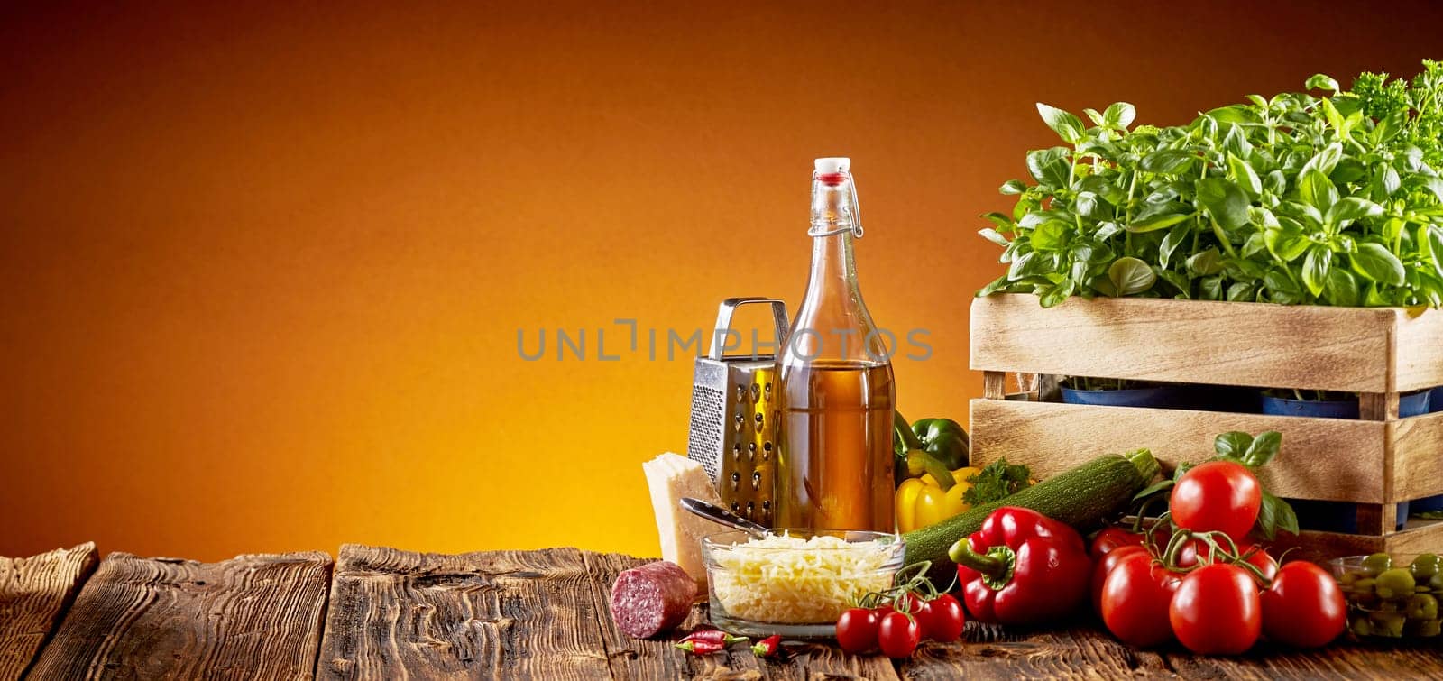 Ingredients for Italian cuisine in a pizzeria with parmesan cheese, olive oil, tomatoes, basil and sweet peppers on a rustic table over a brown background with copy space in a wide angle panorama with glow of the oven fire