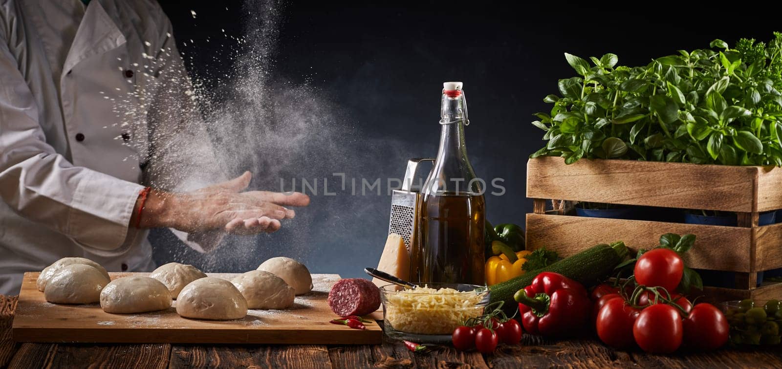 Chef clapping his hands with suspended flour in a freeze motion view as he places portions of raw dough alongside fresh pizza ingredients on a rustic table