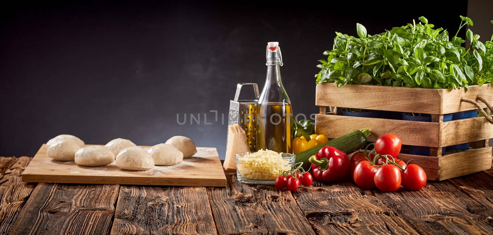 Panorama banner with fresh pizza ingredients, grated cheese, olive oil and mounds of individual portions of dough on a cutting board on a rustic kitchen table over a dark background with copy space