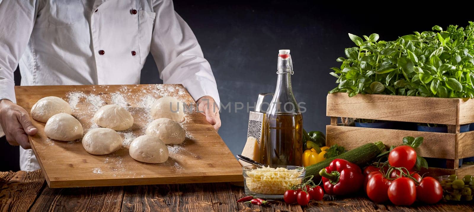 Chef or baker carrying a board with portions of raw dough to a table with an assortment of fresh pizza ingredients in a pizzeria in a panorama banner
