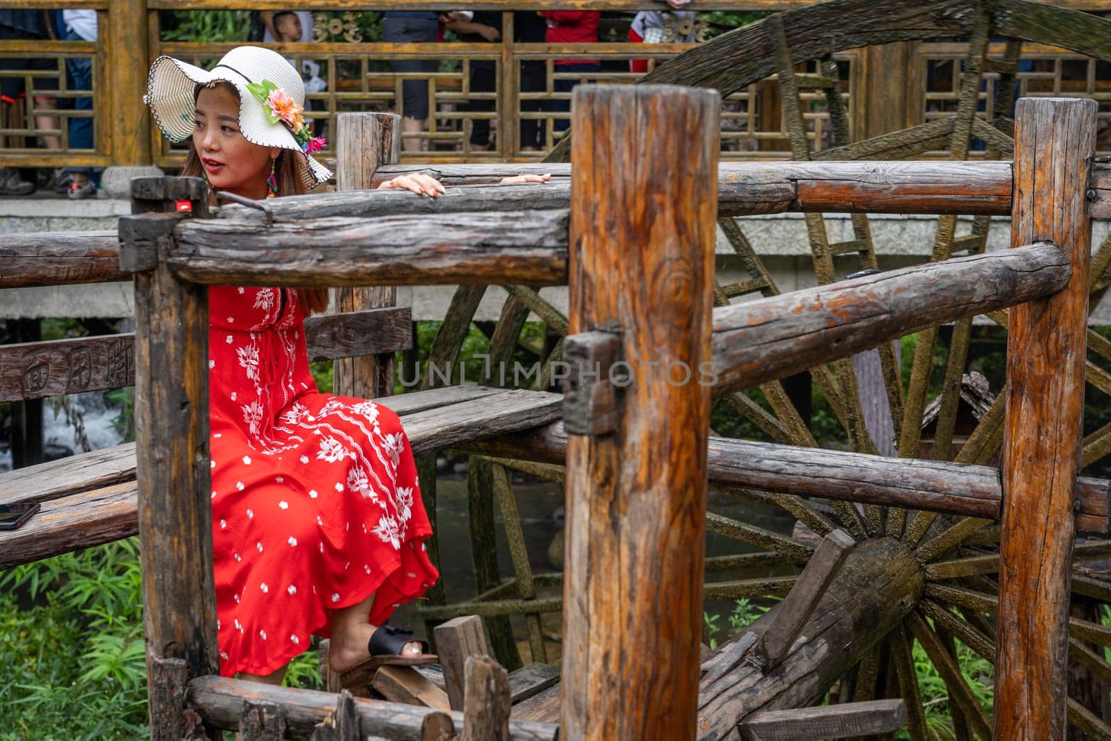 Wullingyuan, China -  August 2019 : Female dressed in a red dress using her legs to pull levers on a wheel of an old Water wheel in Huanglong Yellow Dragon Cave scenic area, Hunan province
