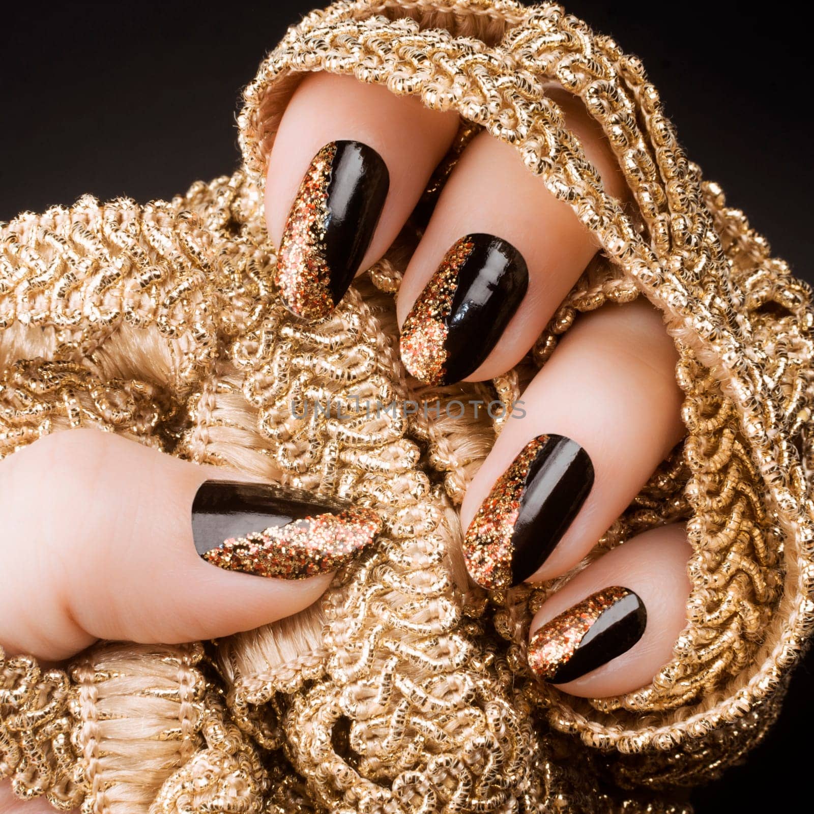 Female hand with golden black nails is holding gold decoration on black background, manicure and nail art concept.