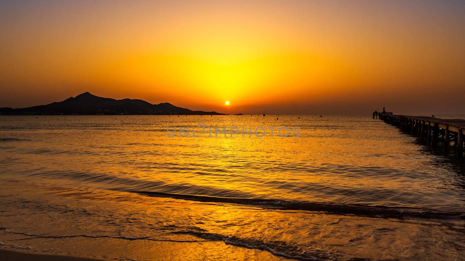 Orange solar disc in sunrise on beach with silhouette of mountains and pier on bay, reach orange and yellow colours of the sky panorama Mallorca Spain