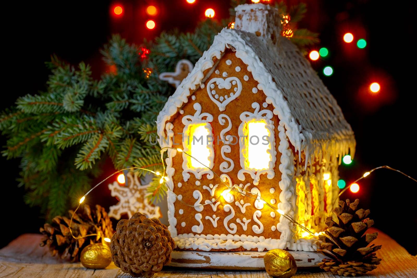 Gingerbread house with bokeh of light bulbs in the background and sprigs of blue spruce