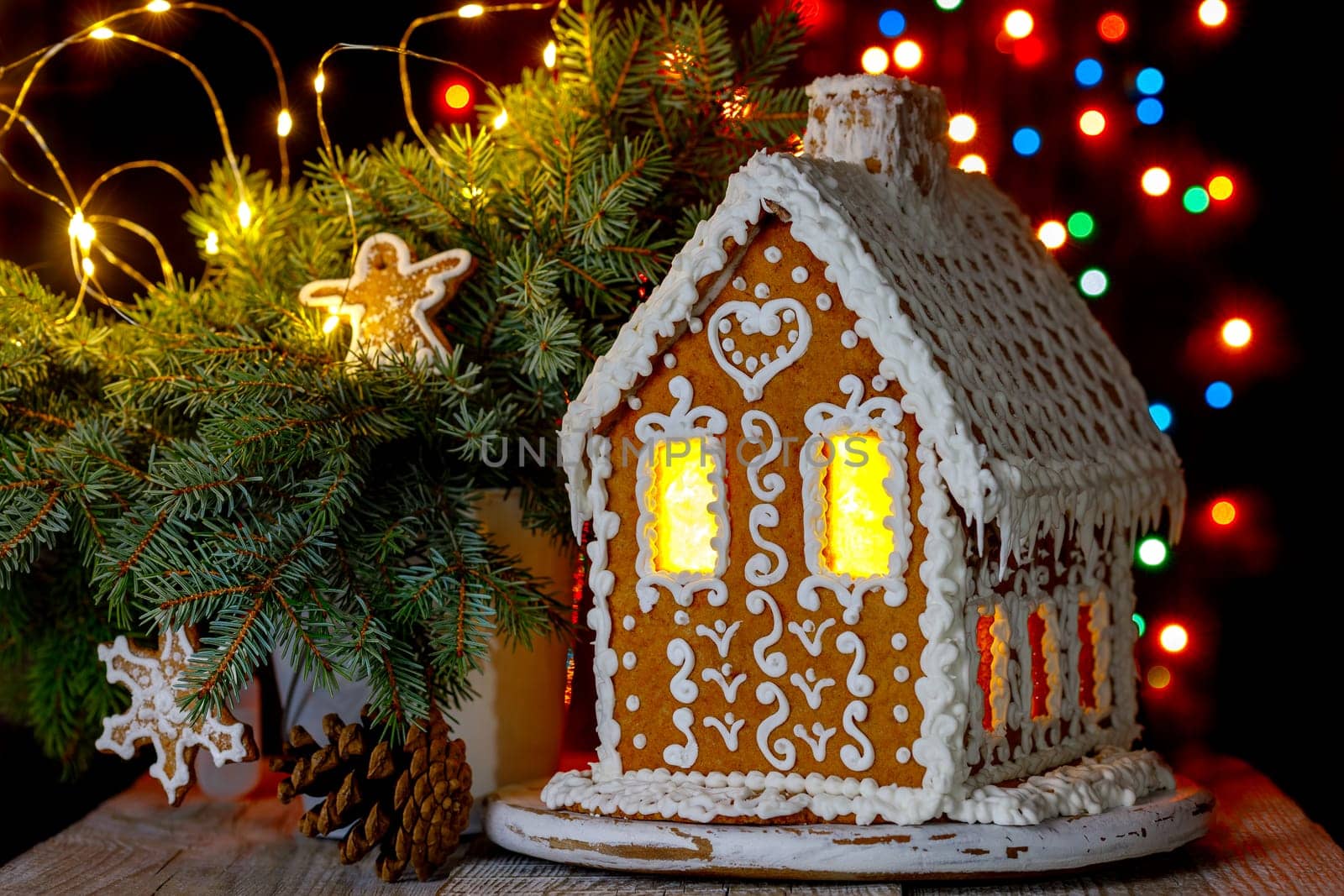 Gingerbread house with bokeh of light bulbs in the background and sprigs of blue spruce