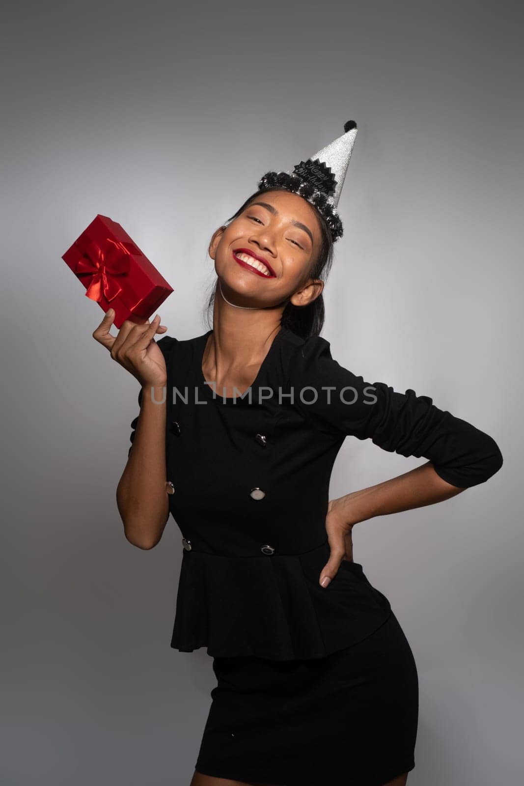 Pretty happy Asian girl in birthday cap holding present isolated over gray wall background