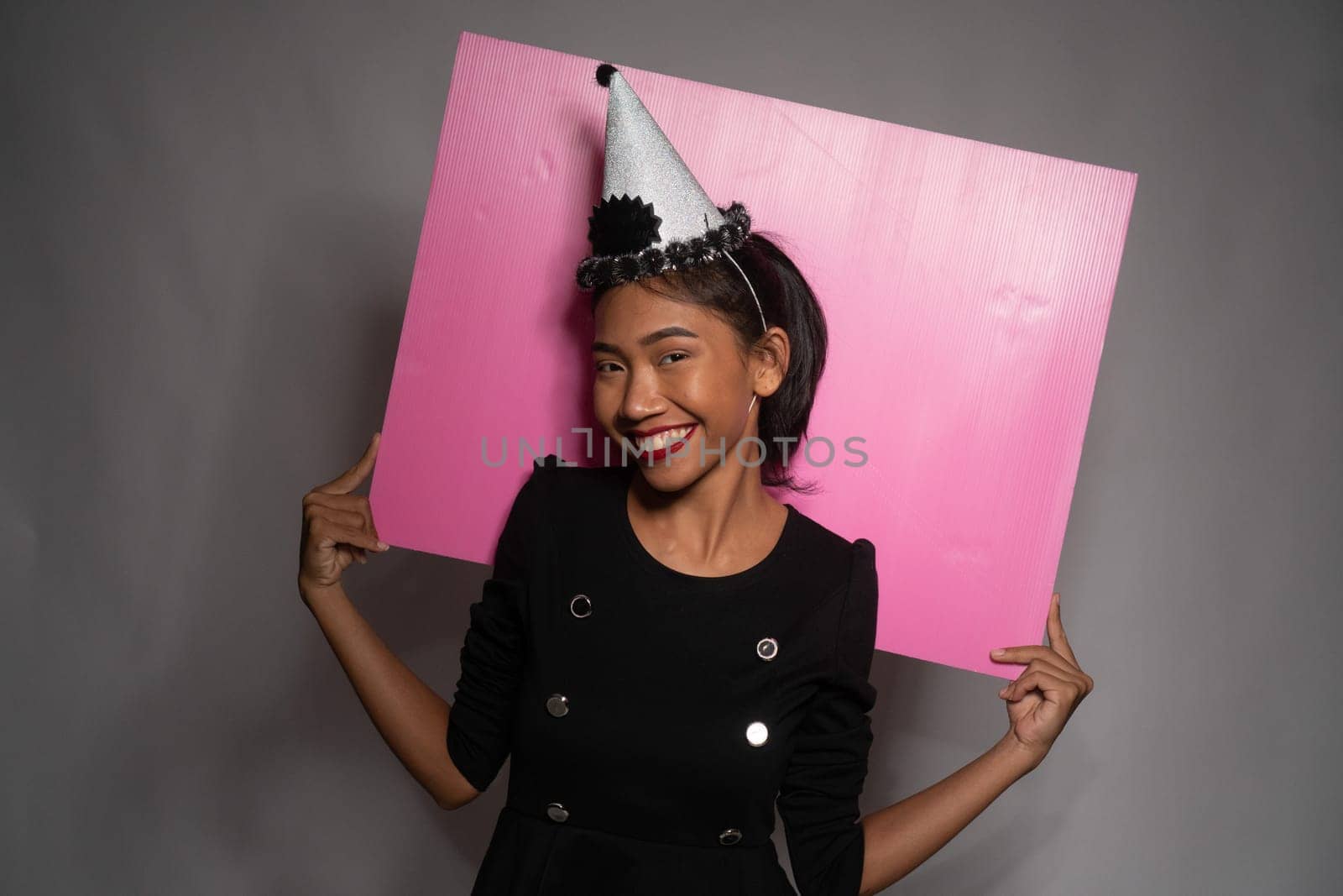 Pretty happy Asian girl in birthday cap holding pink board behind her isolated over gray wall background