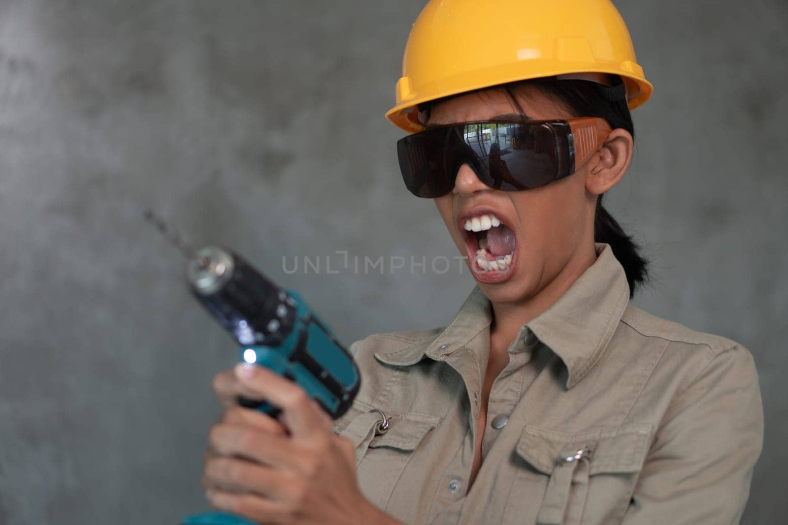 Portrait of attractive young girl builder with drill in overall, helmet and protective glasses isolated over concrete wall background