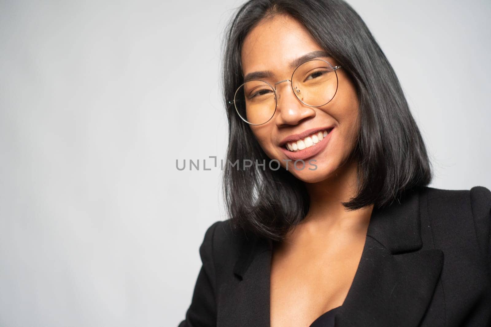 Portrait of attractive young Asian smiling woman in elegant black blazer and glasses posing over light gray wall background. Mock up copy space