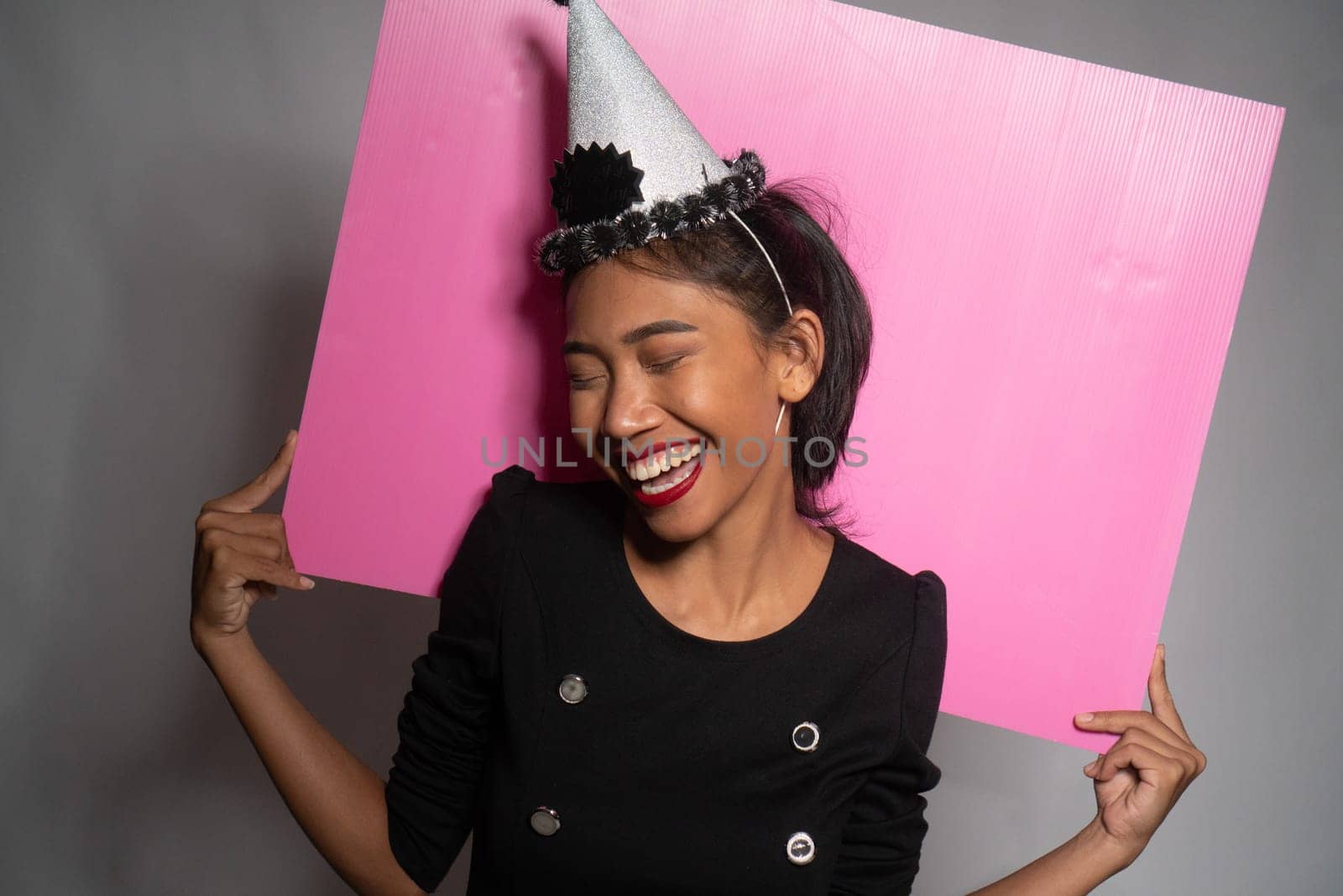 Pretty happy Asian girl in birthday cap holding pink board behind her isolated over gray wall background