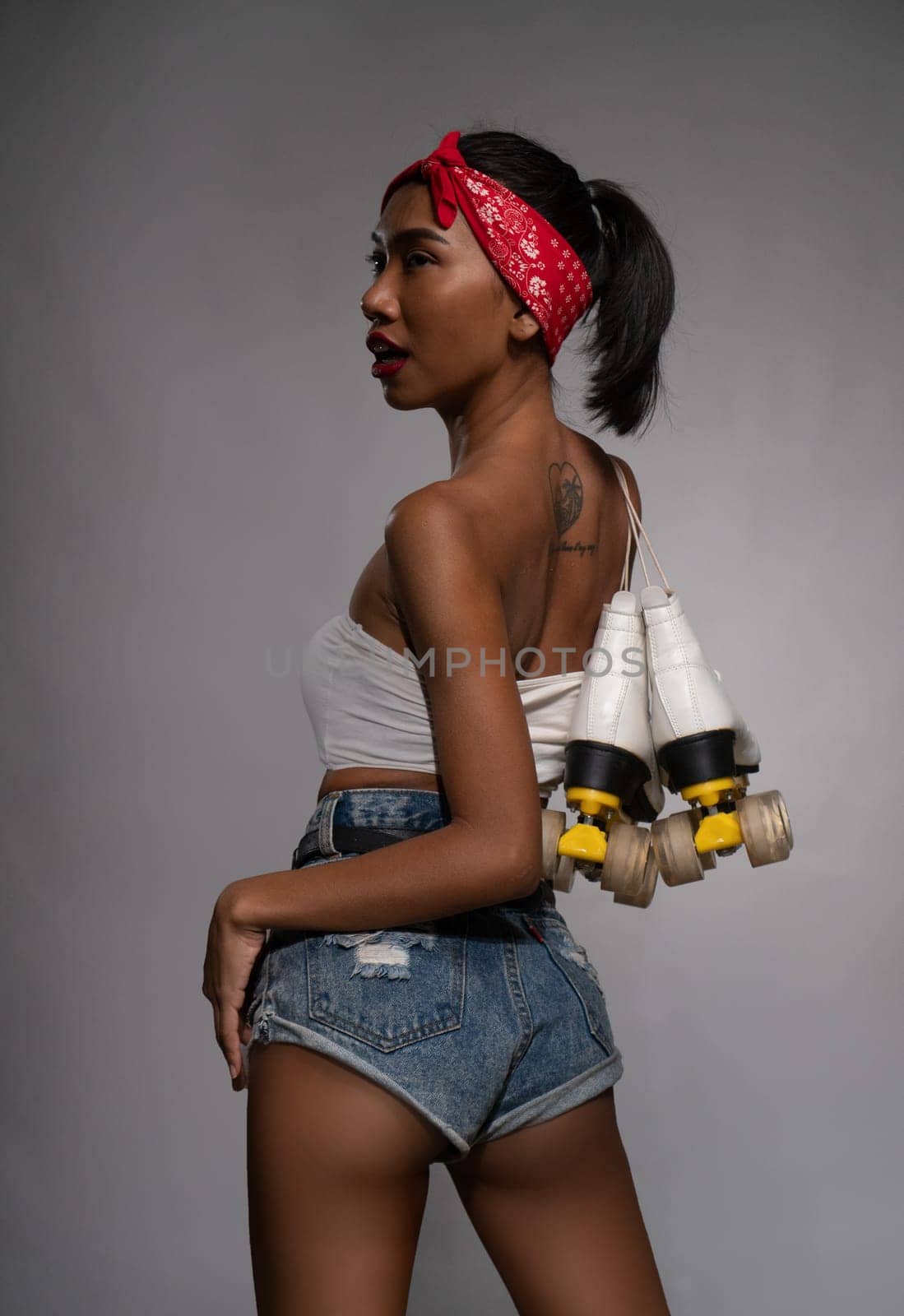 Back view of young beautiful Asian girl holding retro quads roller skates posing in studio over concrete wall background