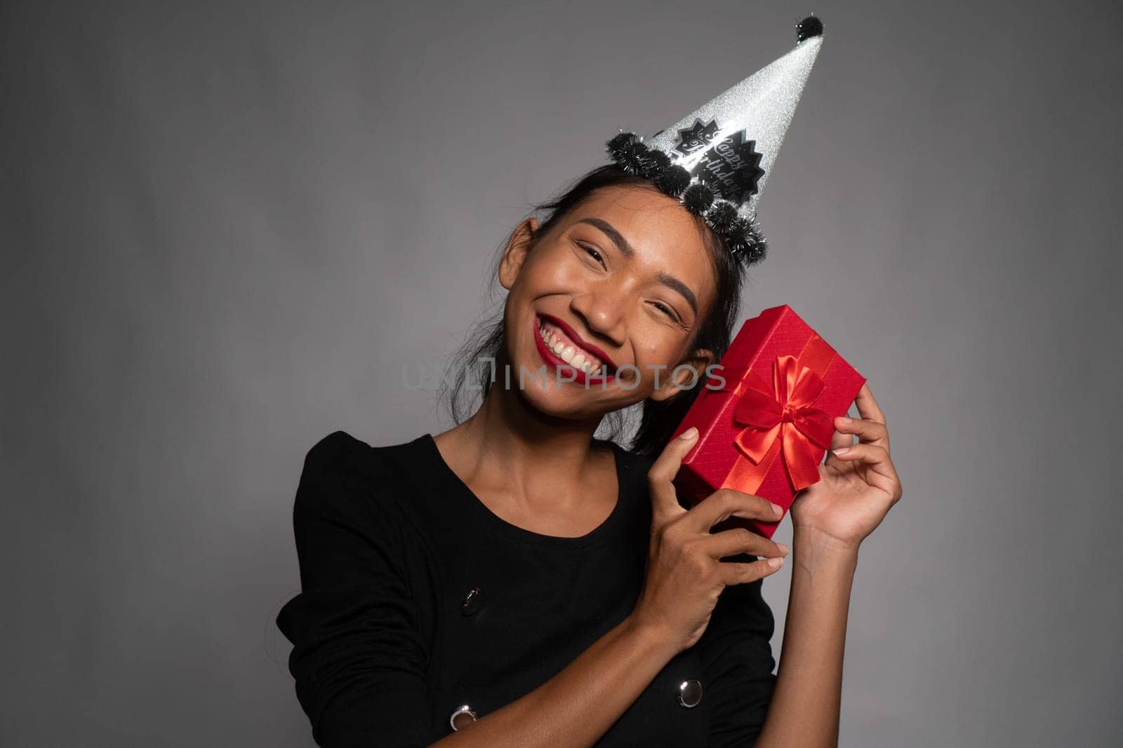 Pretty happy Asian girl in birthday cap holding present isolated over gray wall background