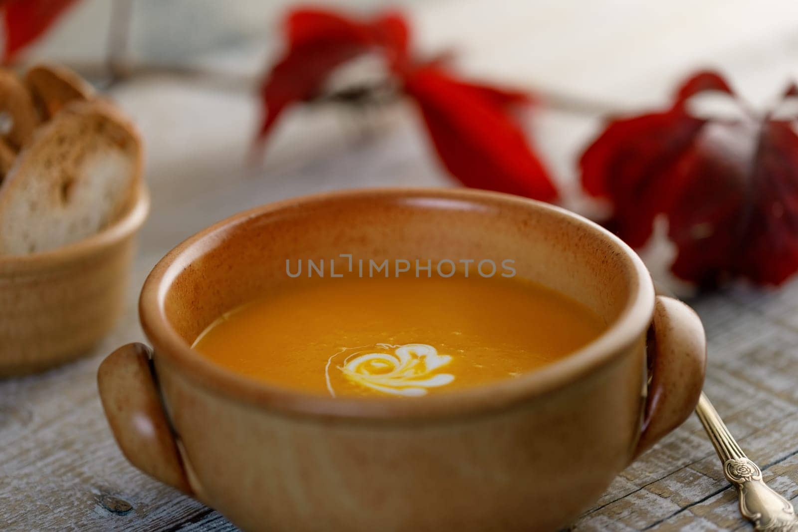 Pumpkin cream soup in a ceramic dish with breadcrumbs and red autumn leaves in the background