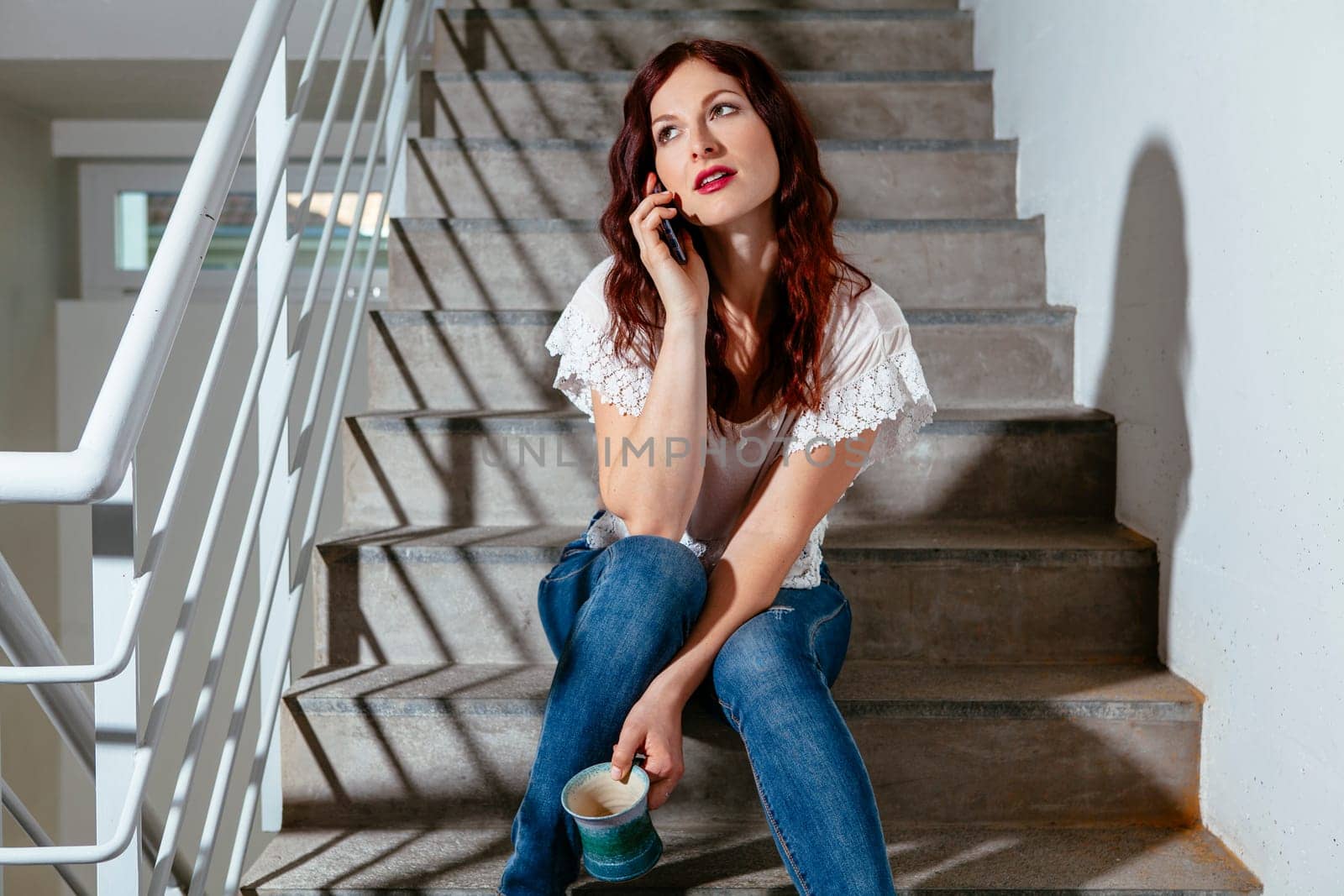 Young woman with red hair sitting on stairs and speaking on her smartphone and drinking coffee.