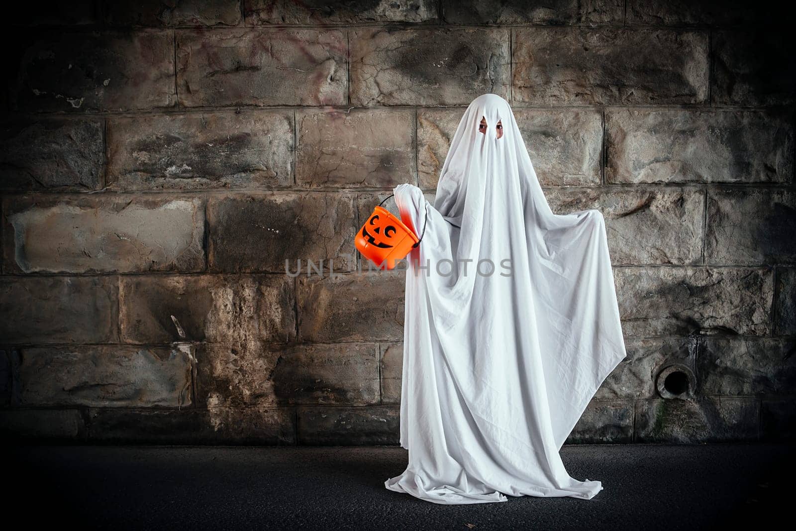 Young boy dressed in a bed sheet to be a ghost, and holding a pumpkin pail for halloween.