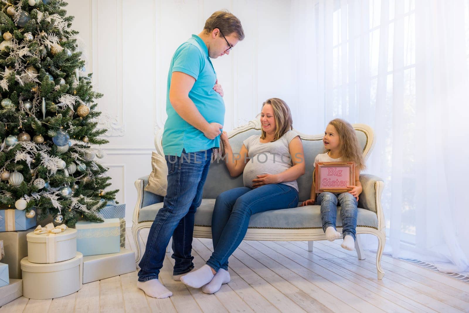 Beautiful pregnant woman sitting with daughter on the sofa near christmas tree, husband standing near