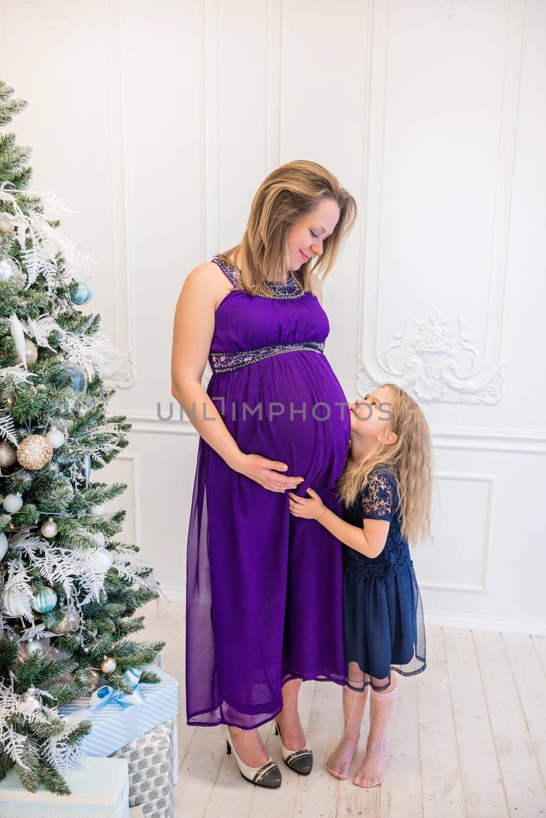 Beautiful pregnant woman in ultra violet dress standing with daughter on the sofa near christmas tree