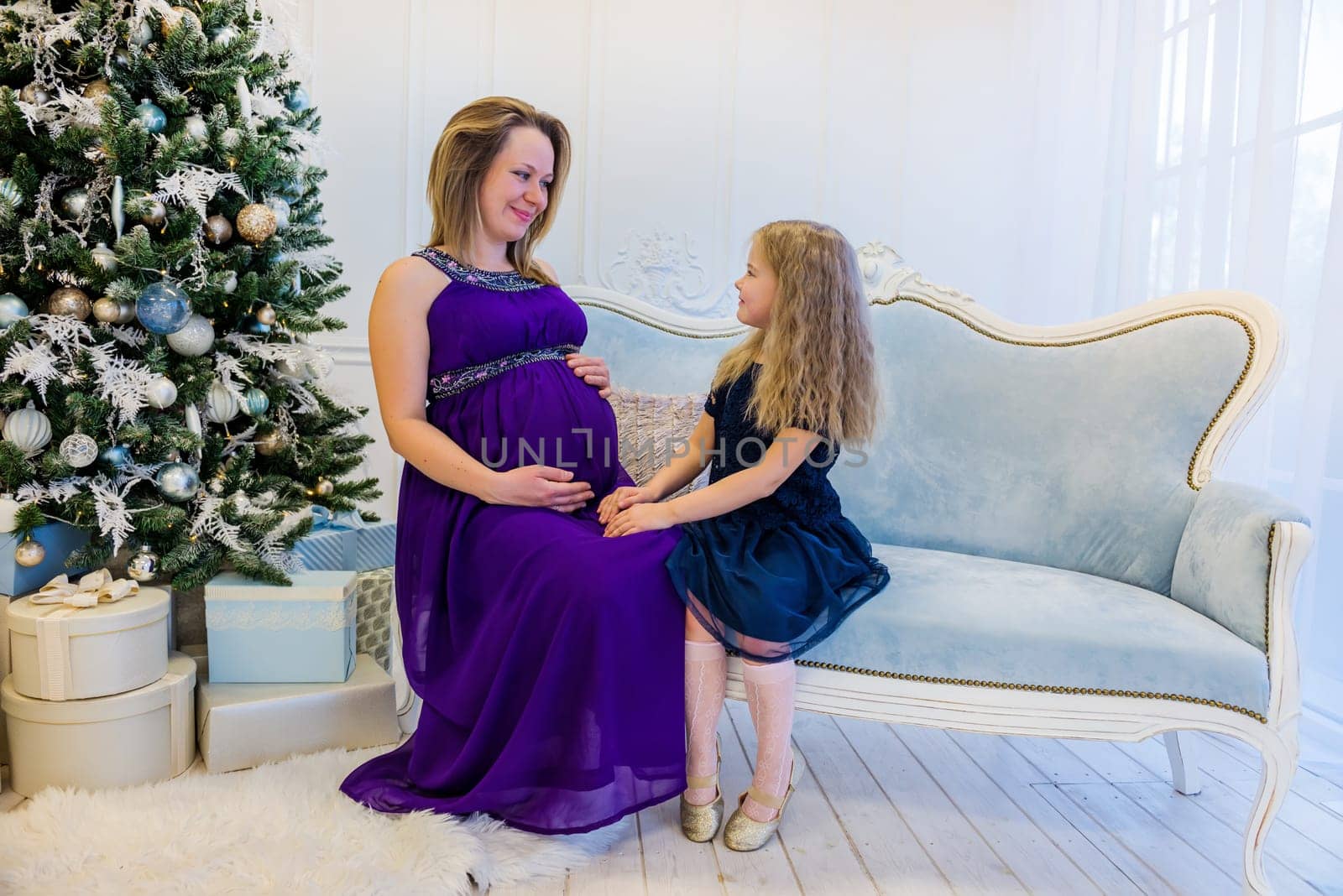 Beautiful pregnant woman in ultra violet dress sitting with daughter on the sofa near christmas tree